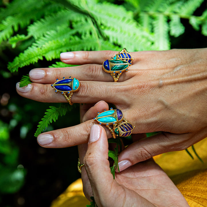 Hands wearing gold beetle rings with blue and green gemstones against a green leafy background