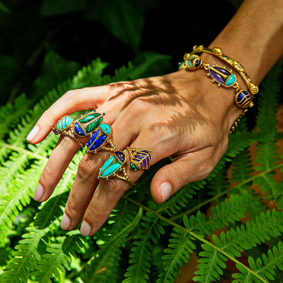 Hand wearing gold beetle rings with blue and green gemstones on a leafy background