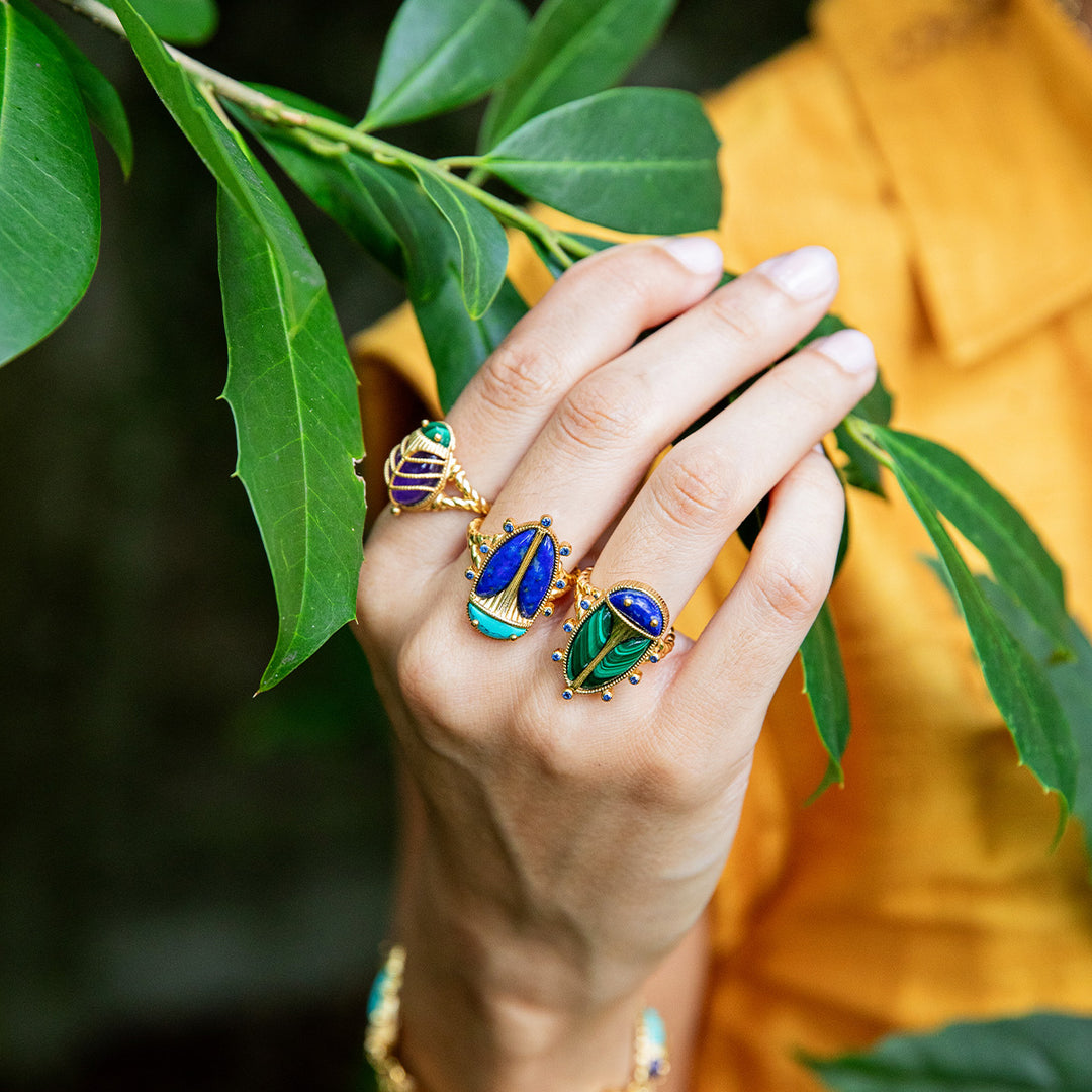 Hand wearing two green and blue beetle rings with green leaves in the background