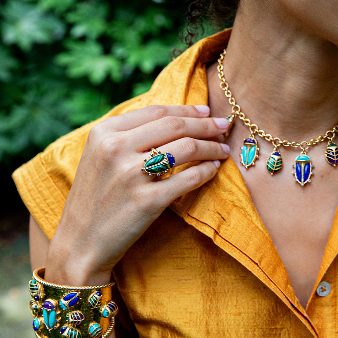 Person wearing gold beetle jewelry with blue and green accents against a blurred green background