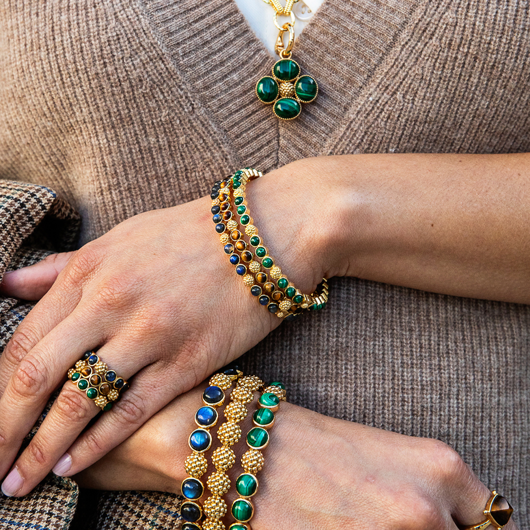 A close-up of a person's hands wearing a coordinated set of gold jewelry with green and blue stones, including a necklace, bracelets, rings, and a watch, styled over a brown ribbed sweater.