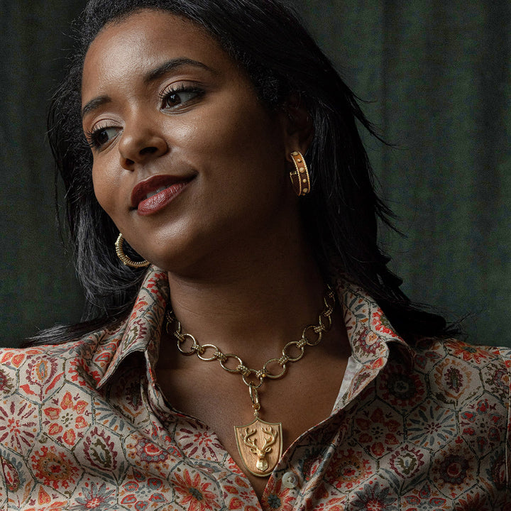 Woman wearing berry and thread gold hoop earrings and gold necklace with a pendant against a dark background