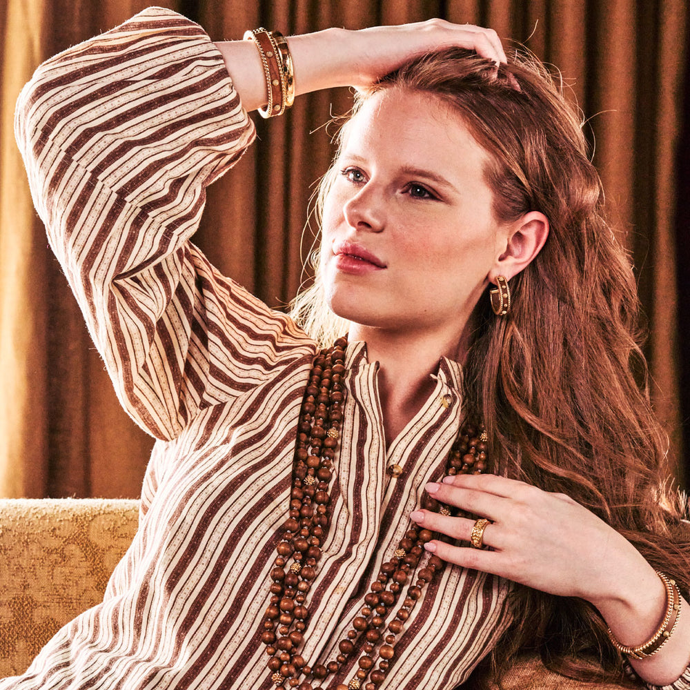 Woman sitting on a couch wearing gold and wooden earrings and bracelets and wooden bead necklaces, with a warm-toned background.