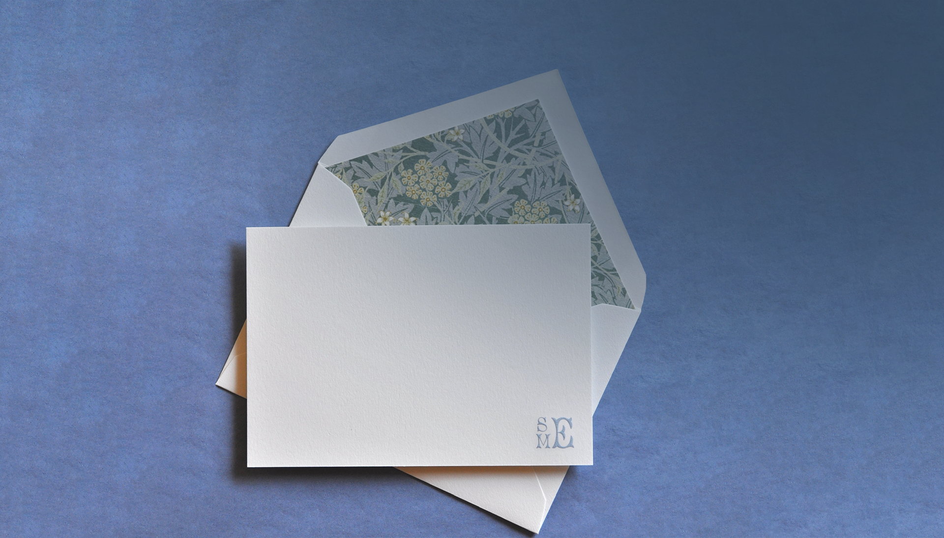 White card with floral envelope liner and cornflower blue engraved monogram  by Arzberger Stationers on a blue background