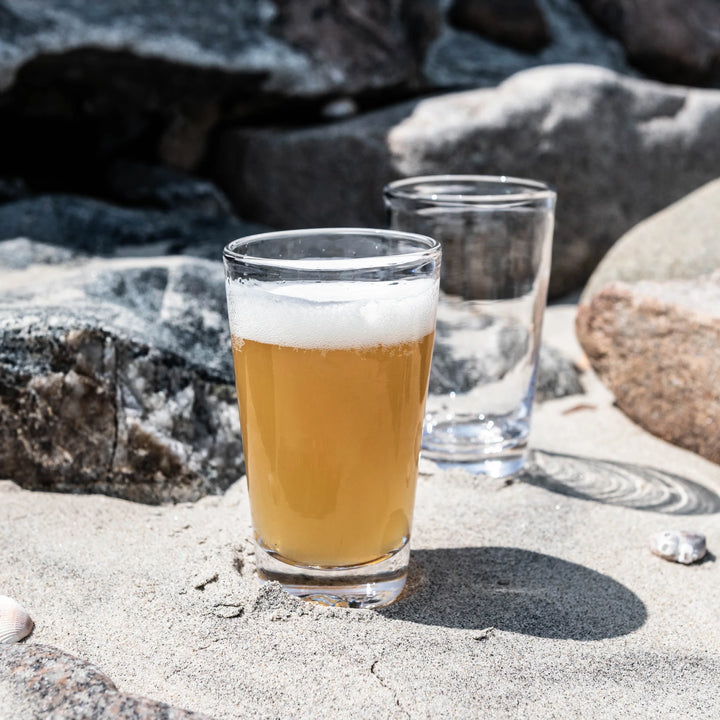 Two glasses of beer on a sandy surface with rocks in the background