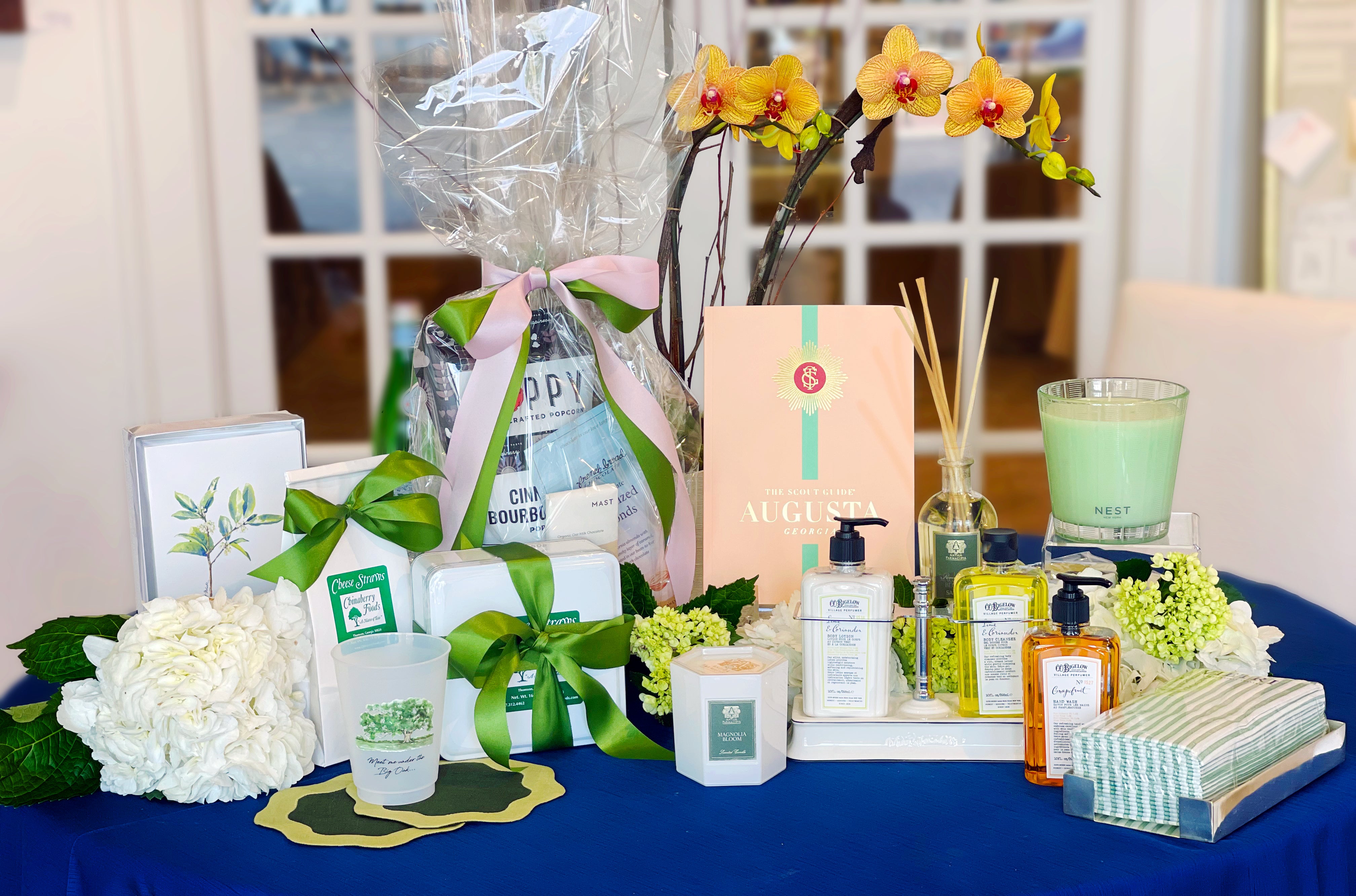 A table with an assortment of gifts including a potted orchid, gourmet snack wrapped in cellophane, a green Nest candle, soaps and lotions from C O Bigelow, A green and white napkin pack in a silver tray, a magnolia bloom candle and reed diffuser, cheese straw packages, a cup with an augusta national theme reading "meet me under the big oak", a card set with a tea olive watercolor by Kat McCall, the Augusta Scout Guide 2026, and many mophead and mini hydrangeas