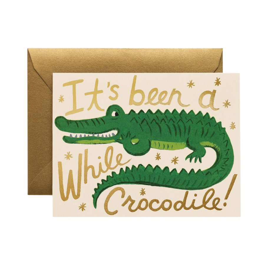 Greeting card with green crocodile illustration and text 'It's been a while crocodile!' ivory paper with a gold envelope.