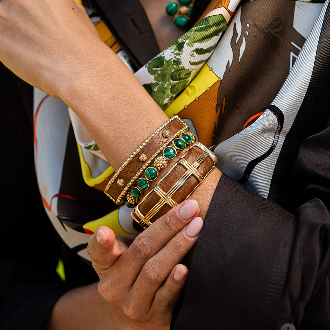Close-up of a hand wearing gold, wooden, and green bangles with a colorful scarf in the background