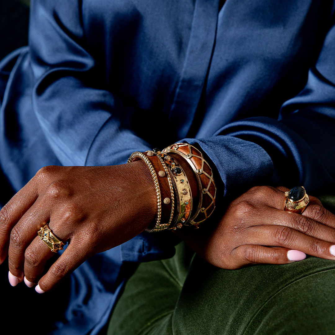 Close-up of a hand wearing gold and wooden bracelets and rings on a blue garment.
