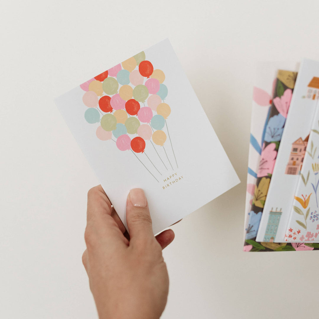 Hand holding a happy birthday card with colorful balloons and other cards in the background on a light surface.