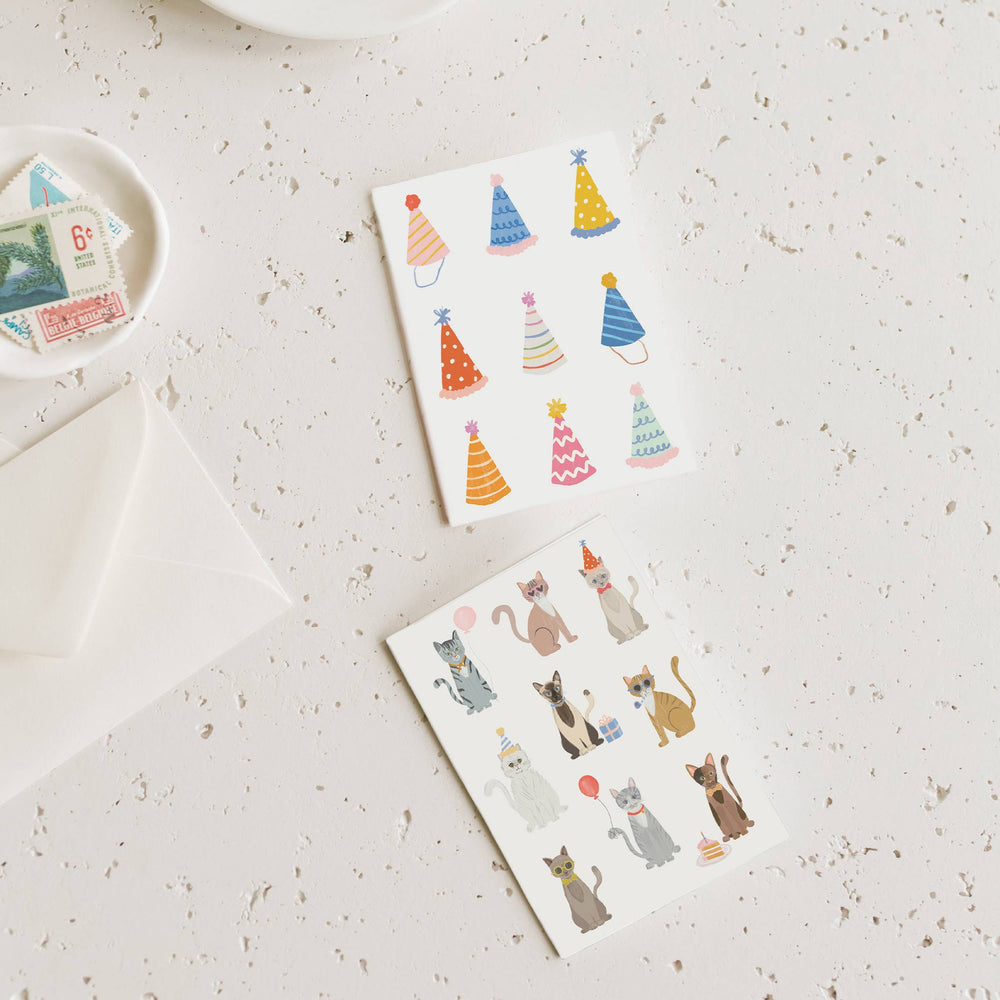 Two birthday cards with colorful party hats and cat illustrations on a textured surface.
