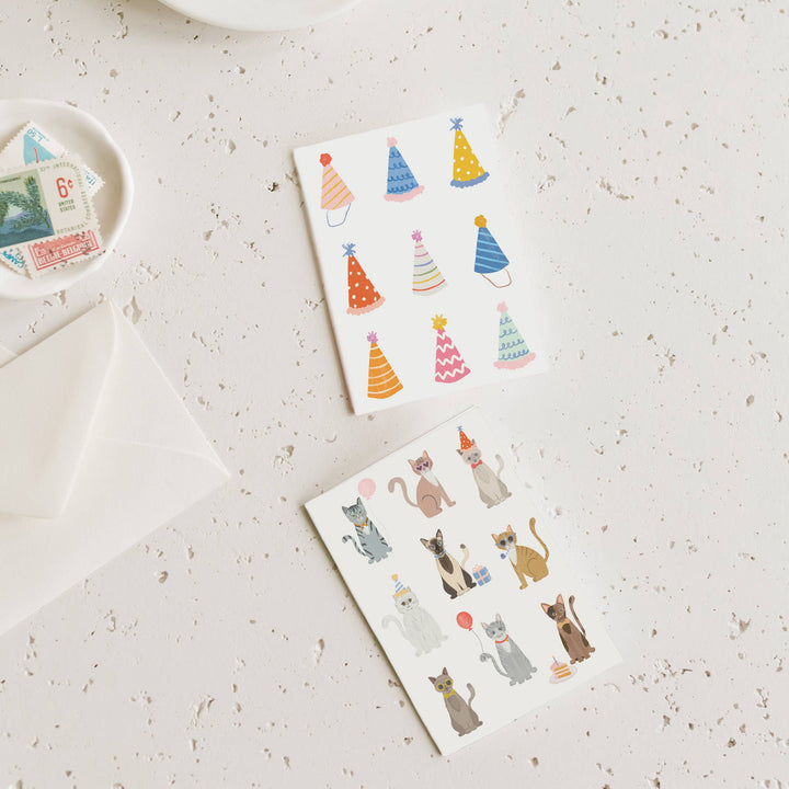 Two birthday cards with colorful party hats and cat illustrations on a textured surface.