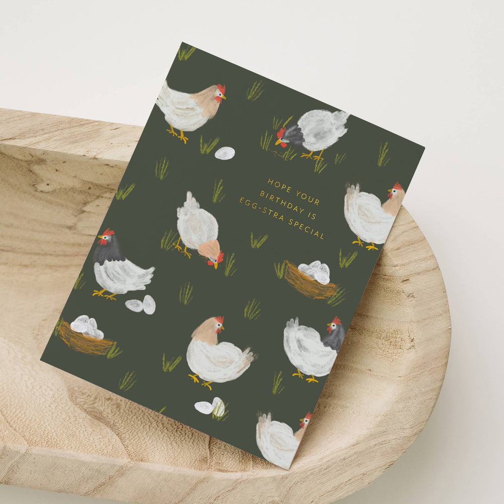 Greeting card with chicken illustrations on a dark green background reading "Hope your birthday is egg-stra special, placed on a wooden surface.