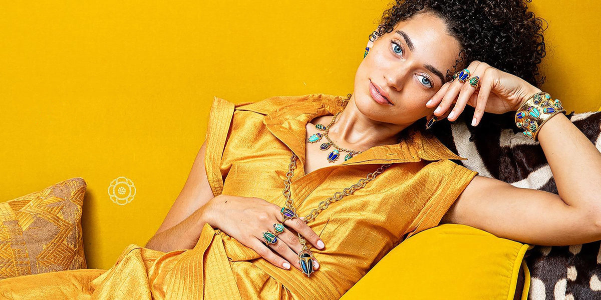 A woman reclining against a bright yellow wall, wearing a shiny mustard-yellow satin dress and multiple colorful statement jewelry pieces with scarab motifs including a necklace, rings, and bracelets.