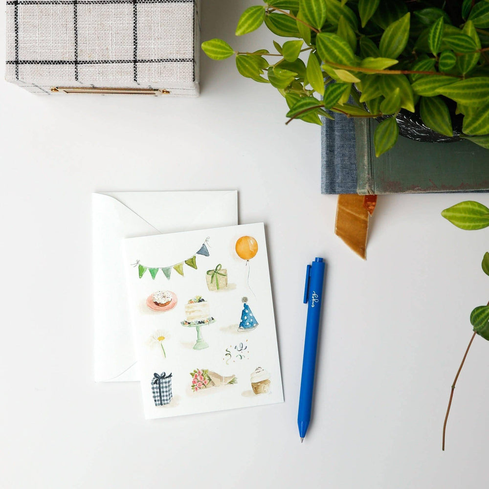 Birthday card with illustrations, blue pen, and decorative box on a white surface with plants.