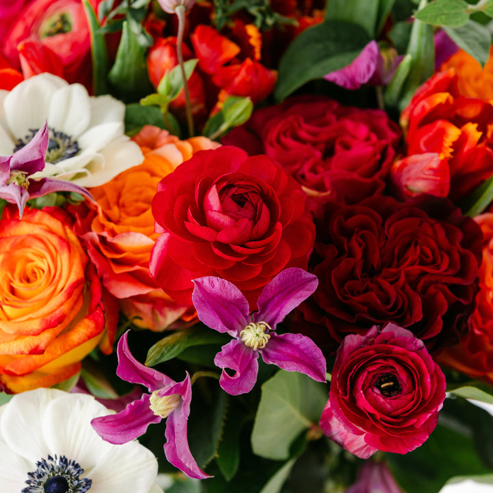 Bouquet of colorful flowers including red, orange, and pink roses, purple clematis, white anemone, with green leaves