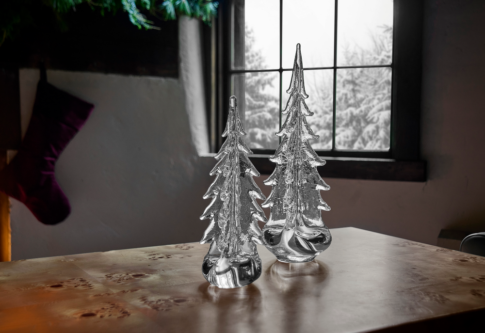Two glass Christmas trees on a wooden surface with a window in the background.