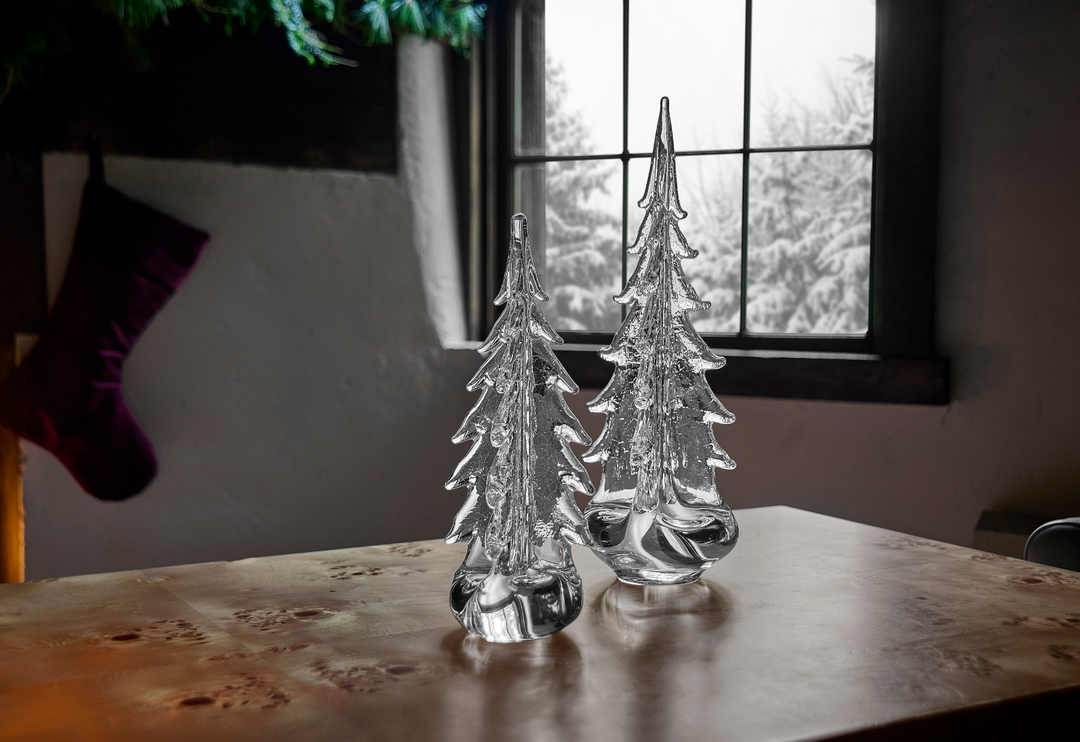 Two glass Christmas trees on a wooden surface with a window in the background.