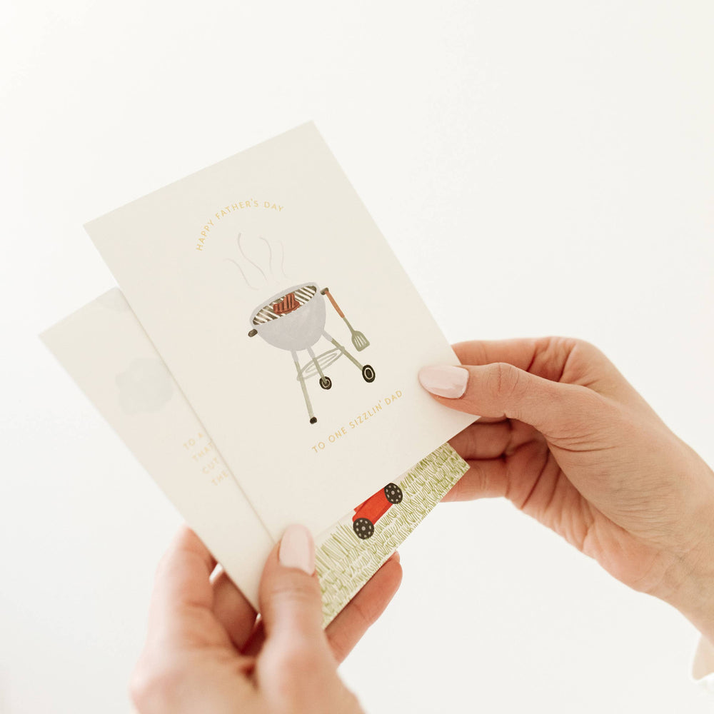 Hands holding two fathers day cards lawnmower and grill illustrations
