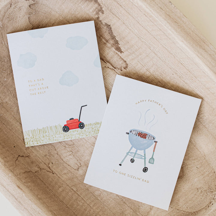Two Father's Day cards with illustrations of a lawnmower and a barbecue on a wooden surface.