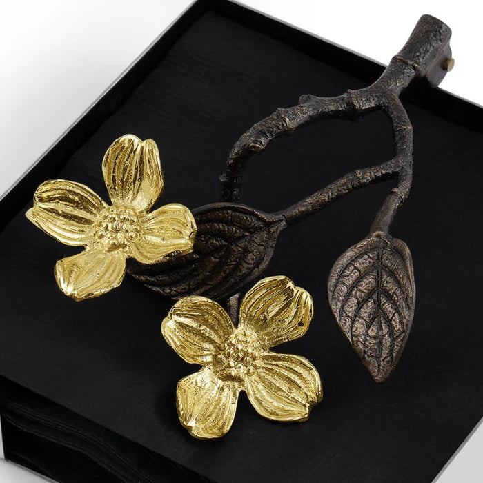Silver square napkin holder with black napkins and a bronze weight shaped like a twig with leaves featuring gold dogwood flowers