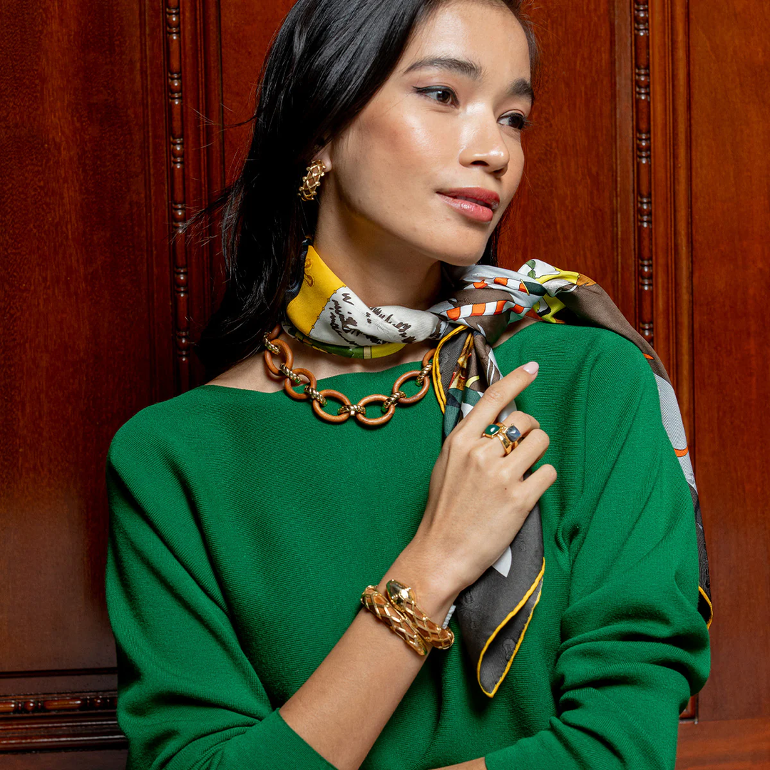 Woman wearing a green top with a colorful scarf, gold wooden chain necklace, and wooden snake bracelet against a wooden background.