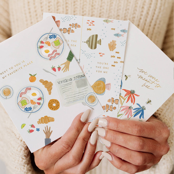 Hands holding a set of colorful greeting cards with various food, flower, and fish designs.