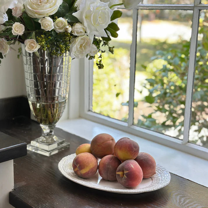 Peaches on a white plate with a lacy rim design in front of a lit window