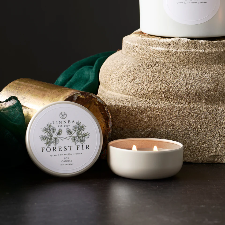 Linnea Forest Fir candle with petit travel tin packaging and a lit candle on a dark surface