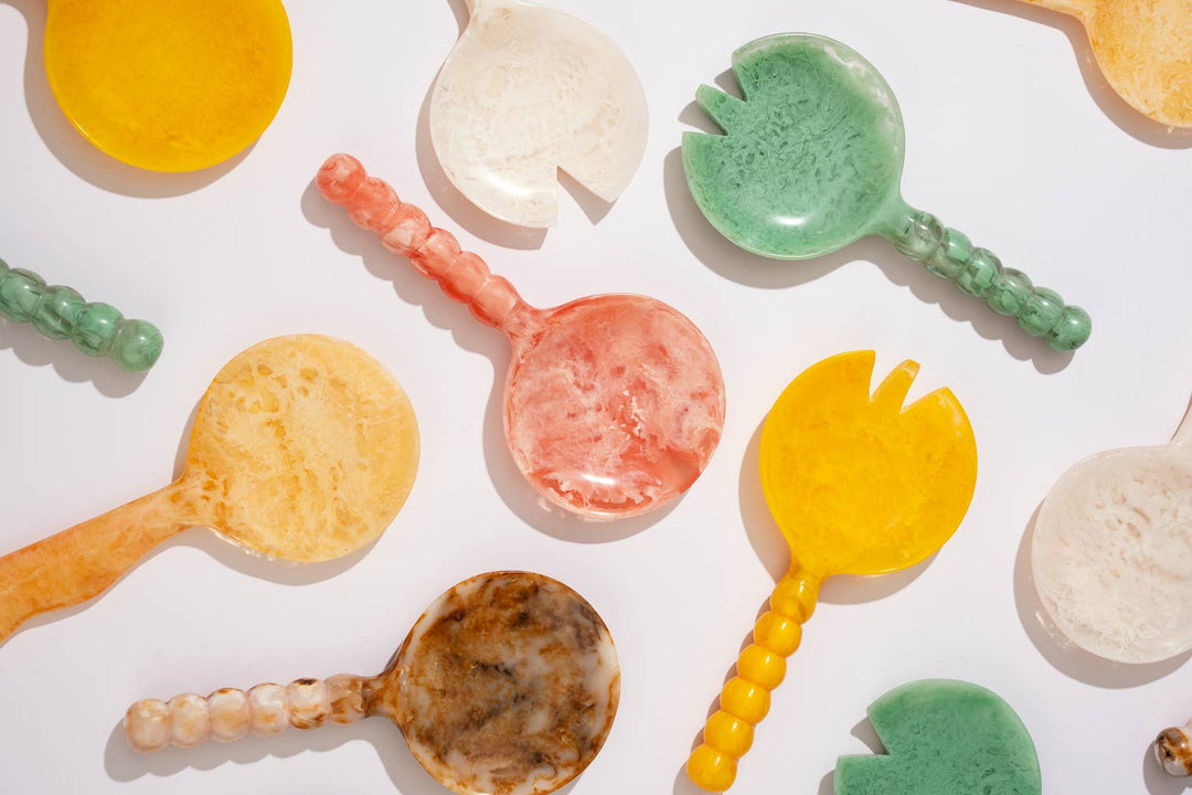 A collection of colorful, handcrafted soap or resin spoons and small dishes scattered on a clean white background. The items feature marbled patterns in yellow, green, pink, brown, and cream.