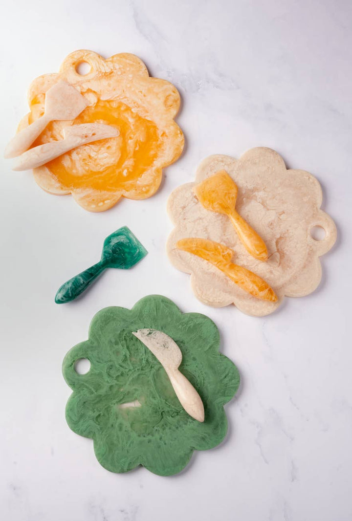 Three scalloped ceramic serving boards in green, beige, and orange, each decorated with a small matching utensil, arranged on a white marble surface.