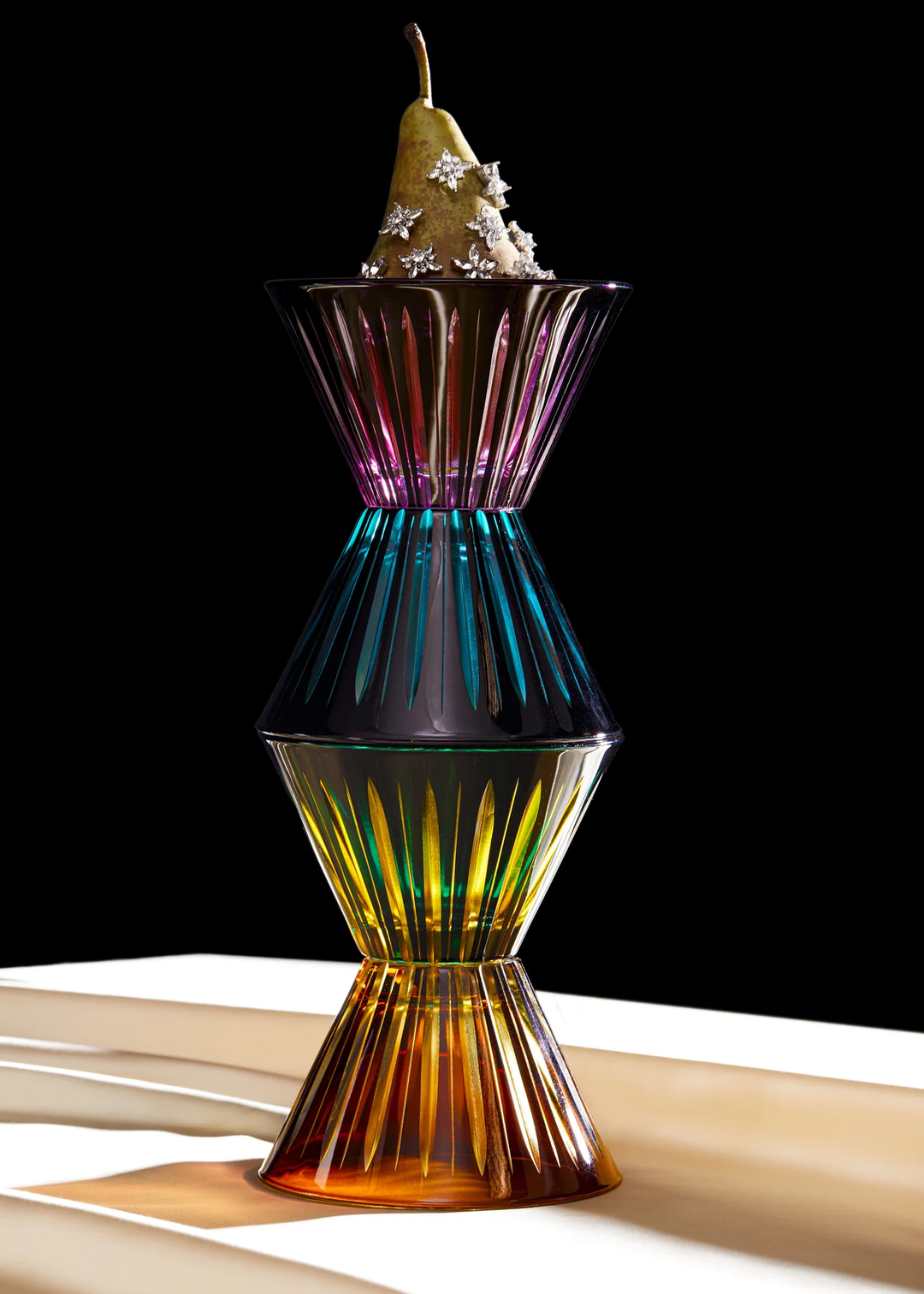 Colorful glass condiments stacked with a pear on top against a black background