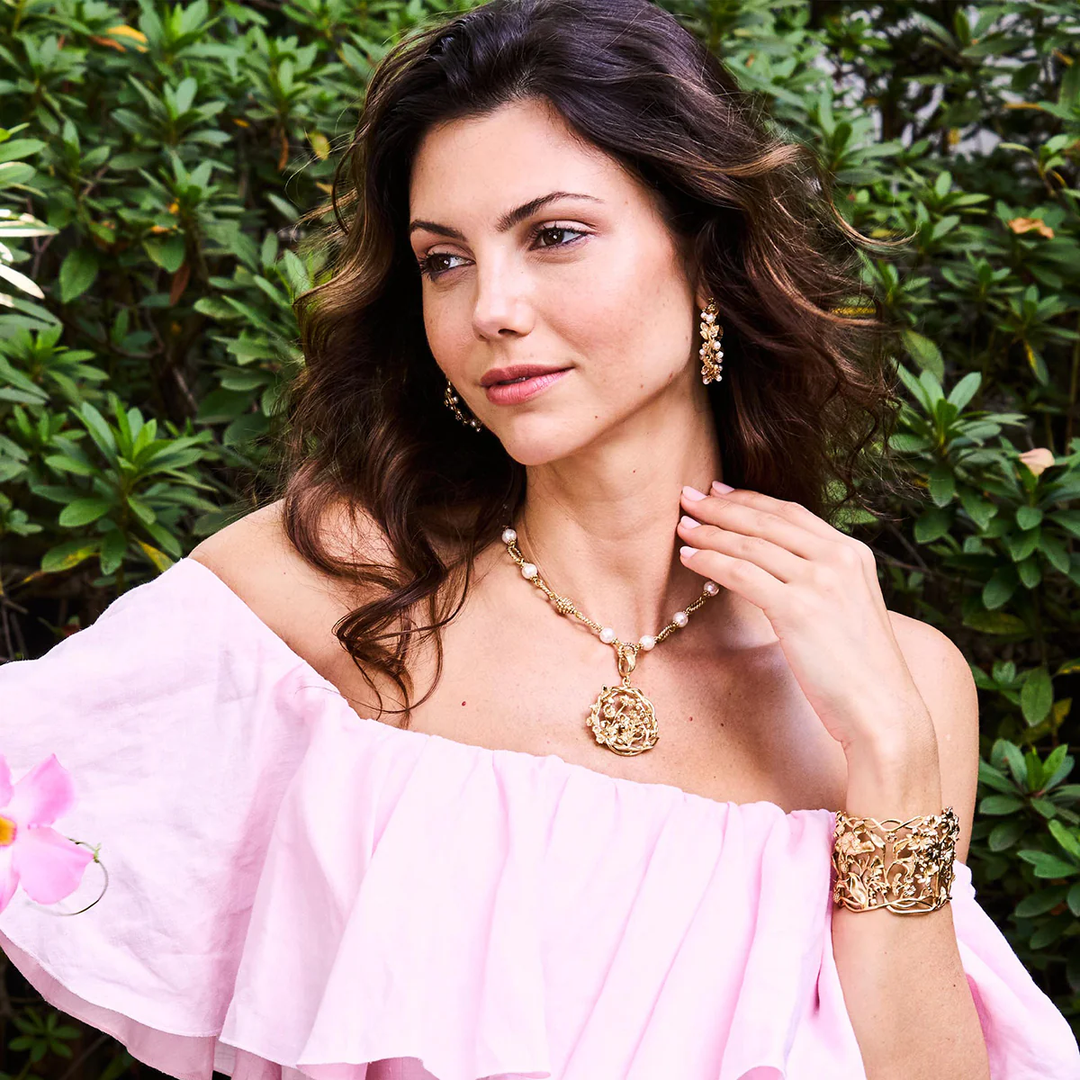 Woman wearing a pink off-shoulder top and elaborate gold floral jewelry against a green leafy background