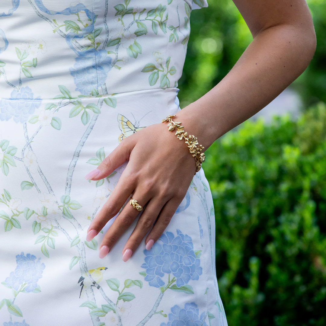 Person wearing a gold floral bracelet and ring with a floral dress against a green background