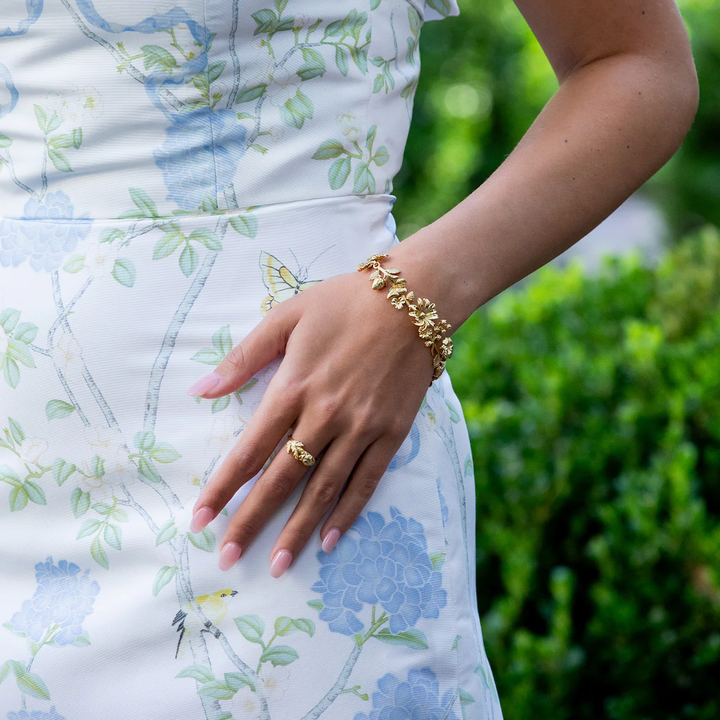 Person wearing a gold floral bracelet and ring with a floral dress against a green background
