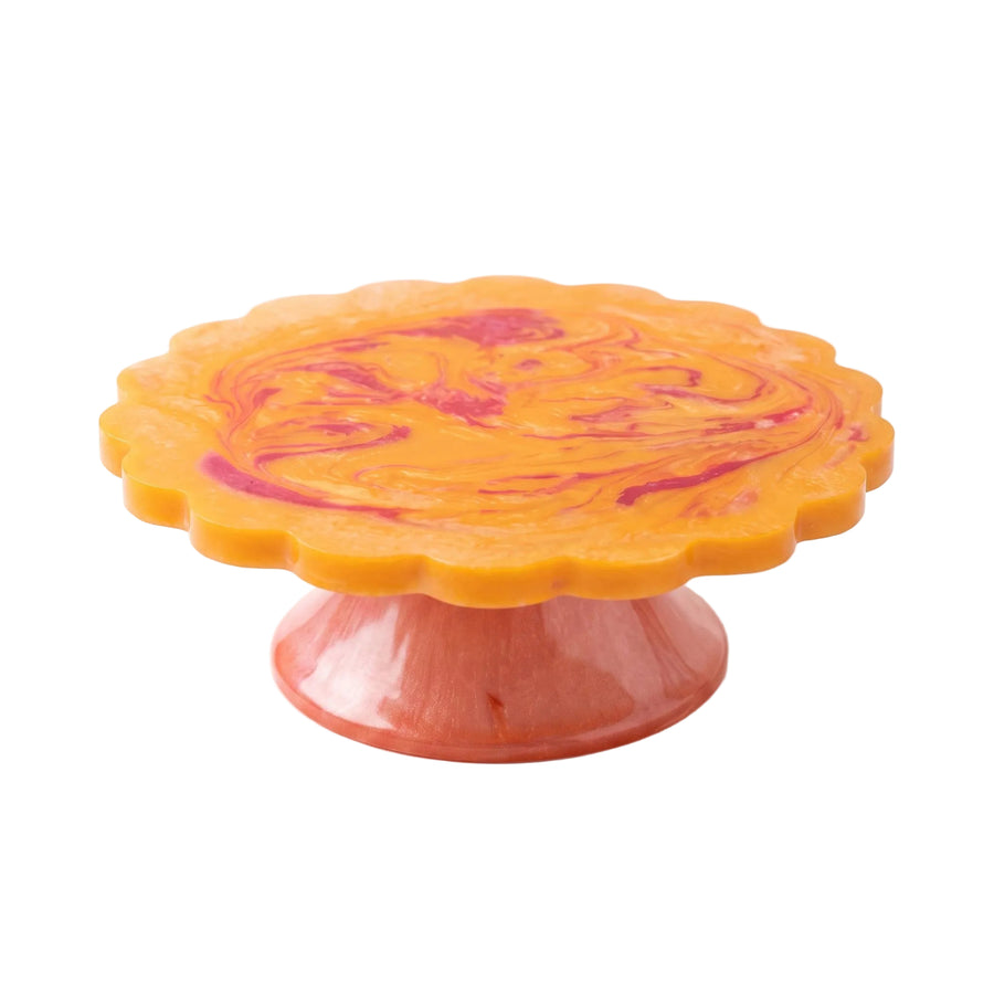 A scalloped cake stand with a marbled orange and magenta top and a pink base, photographed on a clean white background.
