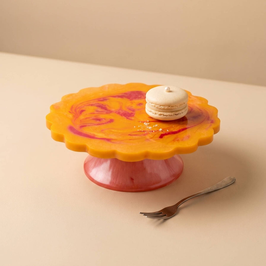 A scalloped yellow cake stand with a pink base and marbled red swirls, holding a small macaron and a fork on a beige surface.