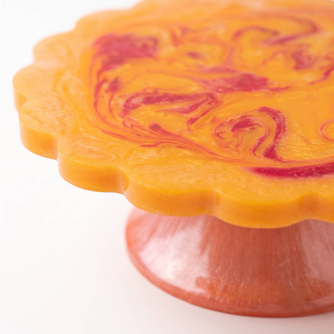 A close-up of a scalloped orange wax melt or soap base with a marbled red swirl pattern on top, supported by a matching orange and red pedestal stand on a white background.