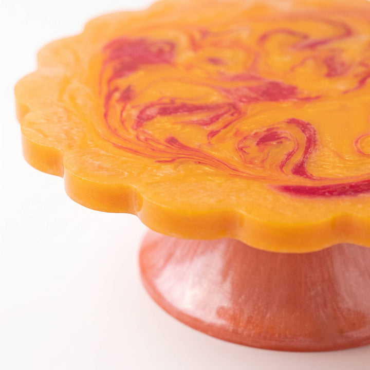 A close-up of a scalloped orange wax melt or soap base with a marbled red swirl pattern on top, supported by a matching orange and red pedestal stand on a white background.