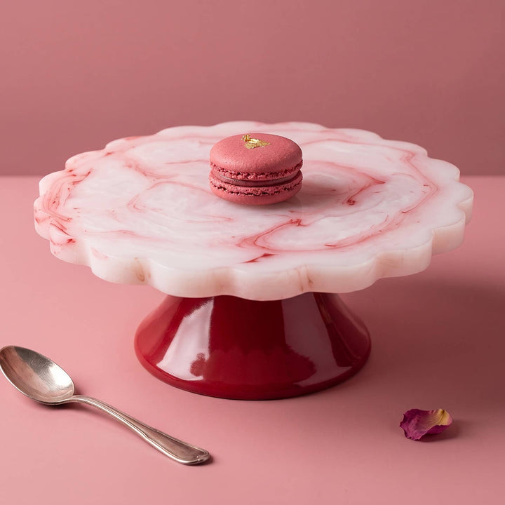 A scalloped white and pink marble cake stand with a glossy deep red base, styled on a pink background with a pink macaron on top, a silver spoon, and a small purple flower petal.