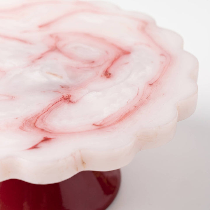 A close-up of a scalloped white top with pink marbled swirls, mounted on a glossy dark red base against a clean white background.