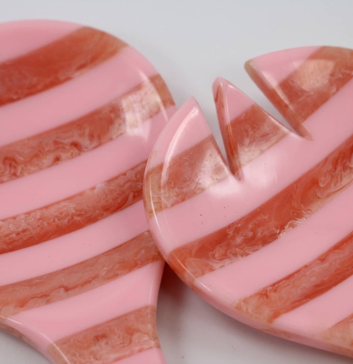 Close-up of two glossy heart-shaped resin beads with alternating pink and peachy-orange marbled stripes on a light gray background.