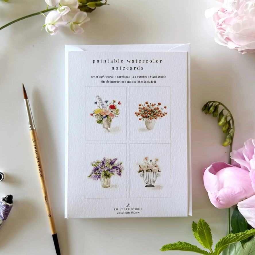 Set of paintable watercolor notecards with floral illustrations on a white surface with flowers and a brush.