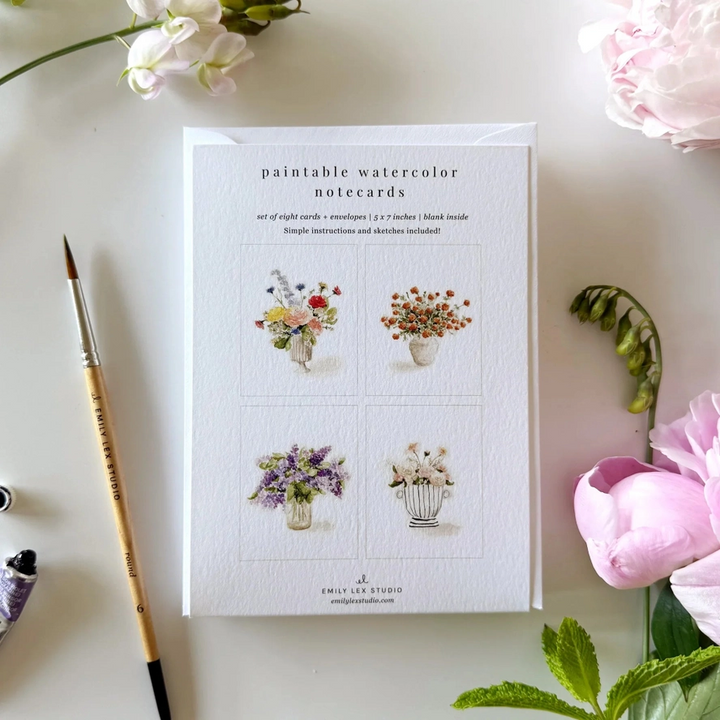 Set of paintable watercolor notecards with floral illustrations on a white surface with flowers and a brush.