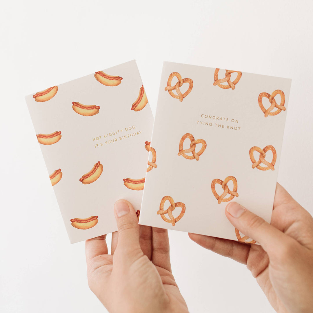 Two small cards with hot dog and pretzel patterns held by hands against a white background