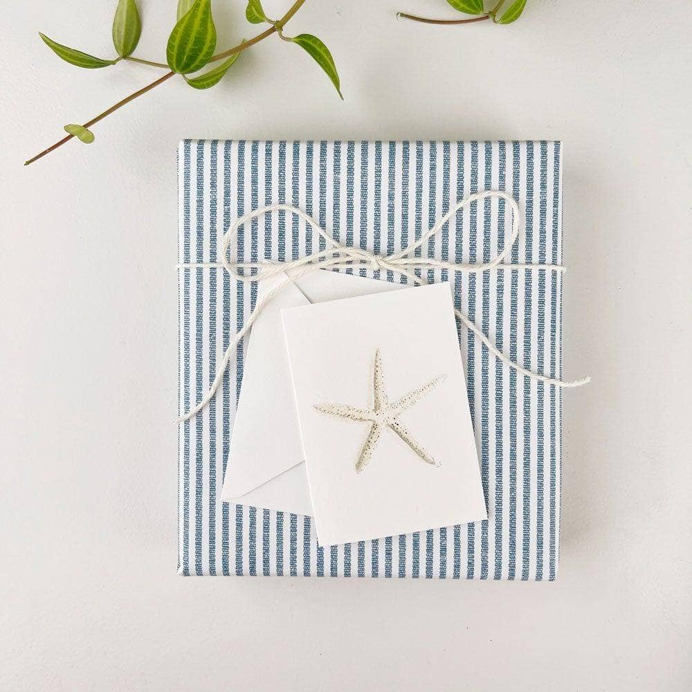 Gift box with blue and white striped paper and a white starfish enclosure card tied onto the front on a light background.