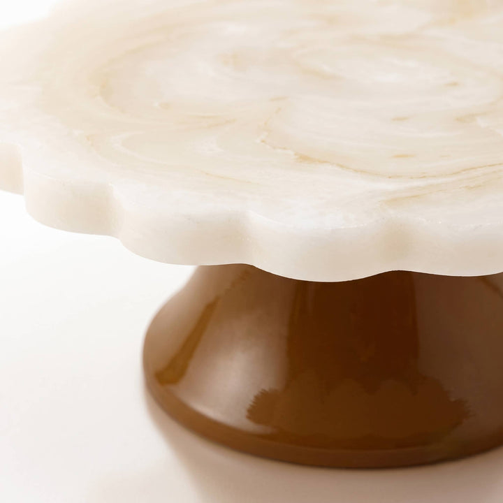 A close-up of a scalloped white stone cake stand with a glossy brown pedestal base on a light background.