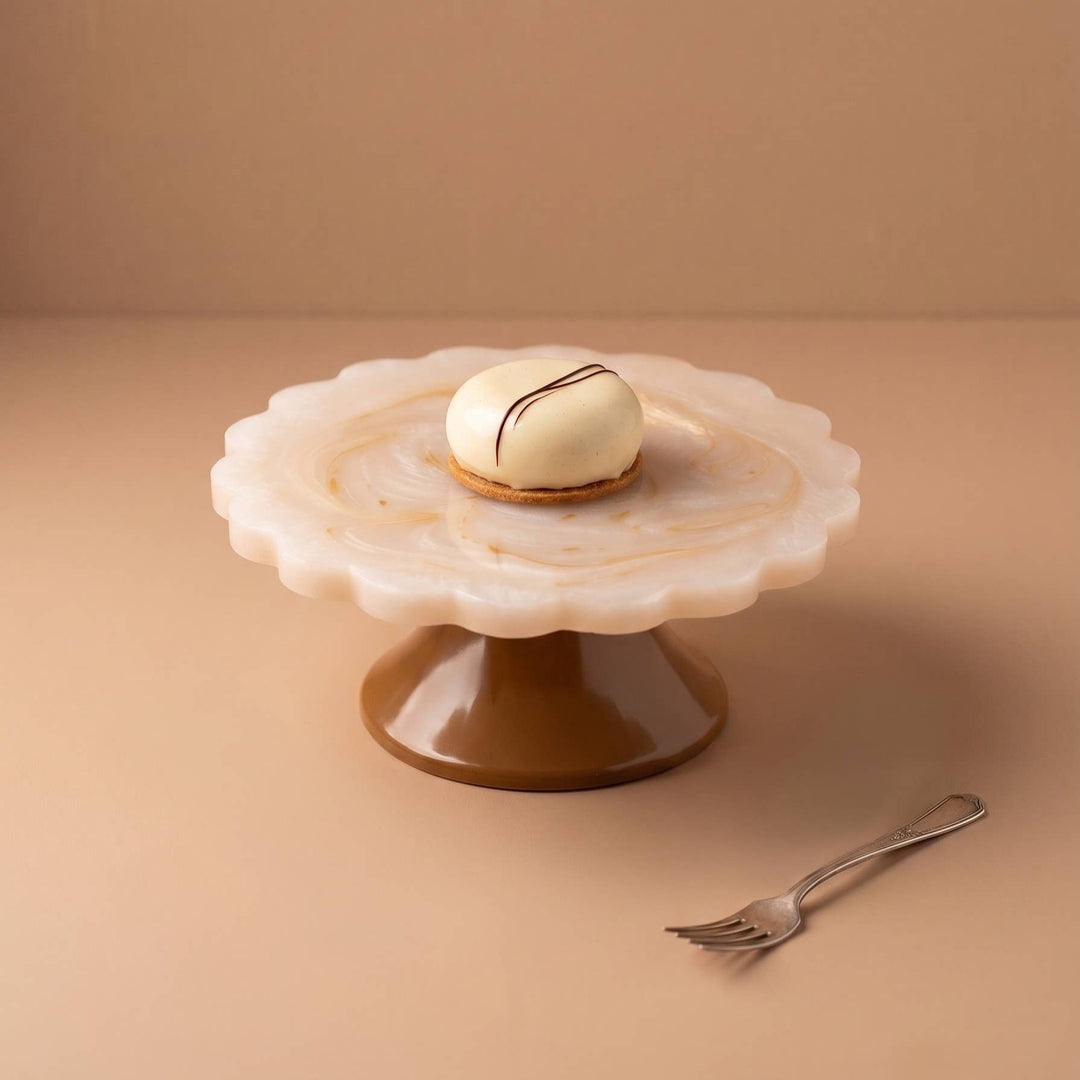A small round white pastry or macaron with a thin dark drizzle sits on a scalloped white cake stand with a brown base, photographed on a warm beige surface with a matching fork nearby.