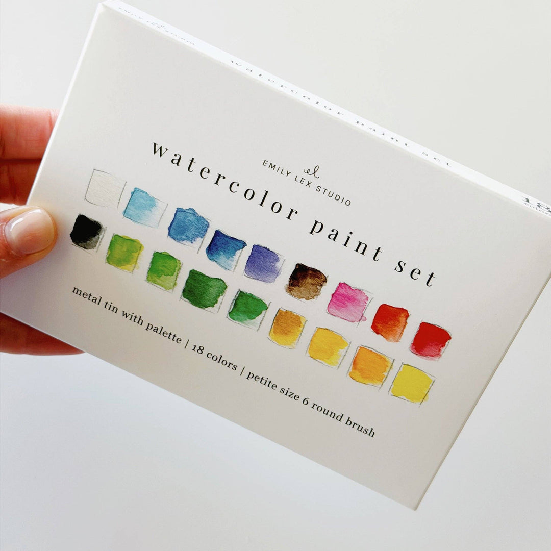 A hand holding a white watercolor paint set box by Emily Lee Studio. 