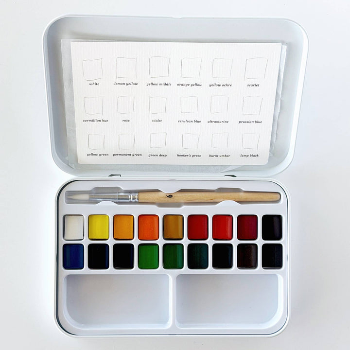 A watercolor paint set in a white metal tin, shown open with 18 color pans, a wooden-handled brush, and a color chart sheet inside the lid 